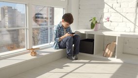 7-8 years old boy sitting at home on windowsill, using mobile cell phone, watching video, playing games. Urban background. 4k slow motion