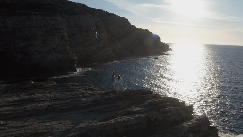 Couple on top of cliff above the ocean enjoy stunning sunset from above. Drone aerial view 