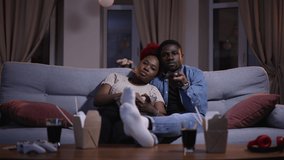 Young african woman, man watching tv and sitting on sofa in home room during lockdown. 4k video American guy hugs female and holds remote control in hand, watches movie and sits on couch in dark