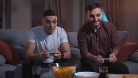 Young men playing game with consoles in hands and sitting on couch in dark living room spbas. 4k video Close-up view of two friends hold digital devices and look with smiles, play and sit on sofa in