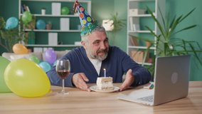 Happy Elderly Man in a Party Cap with a Glass of Wine Celebrates his Birthday Online Using a Laptop, Chatting Cheerfully, Blowing out the Candle on the Cake. Holiday Online, Positive Emotion Concept.
