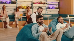 Three co- workers are playing video games and enjoying each-others company while sitting on bean bag chairs, three women behind them are sitting on the sofa watching them play