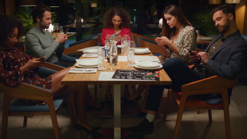 Friends table reading mobile phone browsing internet social media in restaurant. Multiracial group meeting sharing cafe table at evening. Young people ignoring each other in bar. Technology concept. Royalty-Free Stock Footage #1087731659