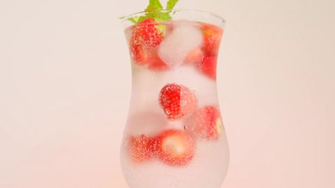 Strawberry drink.summer cocktail.Summer drinks. Mineral water with ice and strawberries in a glass goblet and ripe strawberries with leaves on a light pink background.