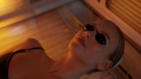 Spa treatments, young female puts on sunglasses, eye protection and sunbathing in a solarium, warm light.