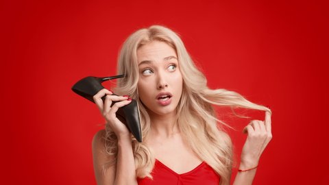 Gorgeus Blonde Female Talking Holding High Heel Shoe Like Telephone Near Ear, Flirting And Communicating Posing In Studio On Red Background. Mobile Communication And Flirt Concept