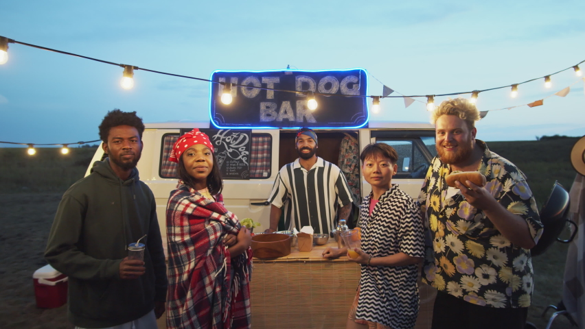 Group portrait of cheerful multiethnic friends and food truck seller smiling and posing for camera with drinks and hot dog on summer festival