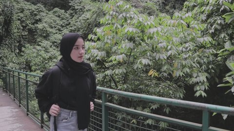 a girl in a black jacket and hijab is walking in the middle of the forest and stops for a moment