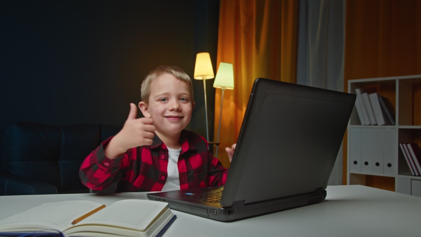 Kid boy is studing at home typing on laptop then looking at camera and showes thumb up