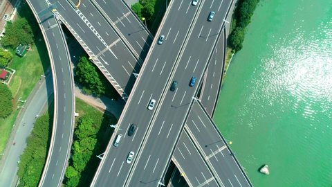 Aerial top down view of a highway overpass multilevel junction with fast moving cars surrounded by green trees and with a river on a side on a sunny day.