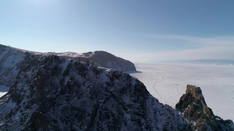 Aerial tilt shot of cape Khoboy, Olkhon island. Tall rocks in frozen lake Baikal with many people and cars around. Popular touristic destination. Winter landscape. Panoramic view