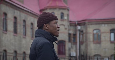 African American man turns to camera smiling widely against ancient building. Black guy in warm beanie and jacket stands on street in old town closeup