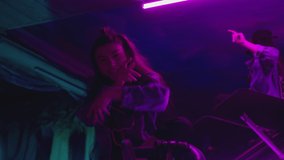 Group of stylish young people dancing , moving near military tank inside garage building Colorful stylish clothes , hippie style fashion . Modern professional dances . Purple led light , slow motion