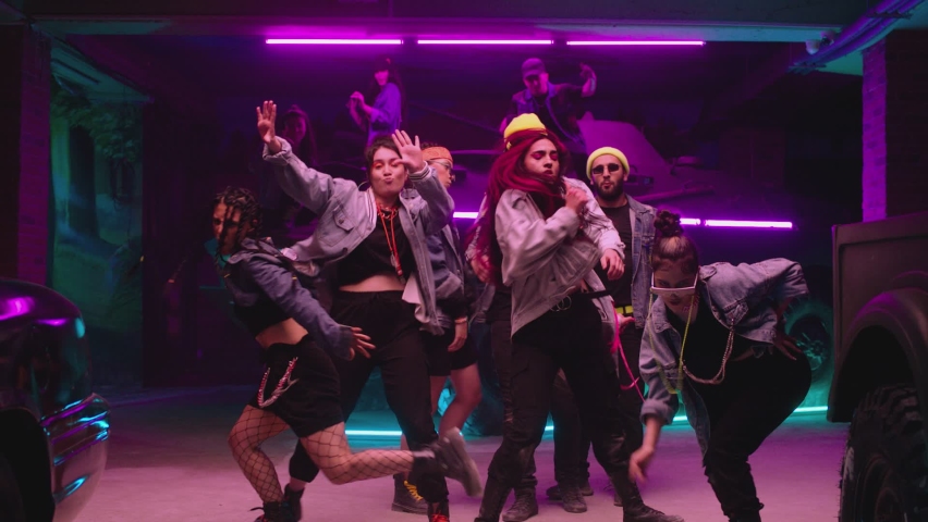 Group of stylish young people dancing , moving near military tank inside garage building Colorful stylish clothes , hippie style fashion . Modern professional dances . Purple led light , slow motion Royalty-Free Stock Footage #1087756322