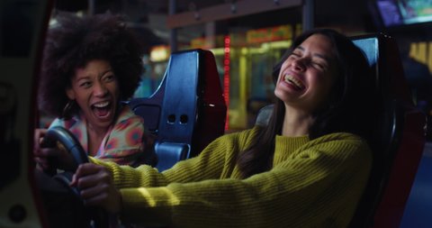 Cinematic shot of young happy interracial girlfriends steering wheel racing in sports car while having fun to play simulation race game together in amusement park on weekend.