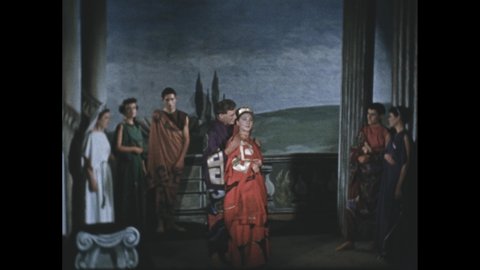 1600s: Actors on stage, man and woman in center speaking. Woman with clapboard. Man and woman in period costume speaking.