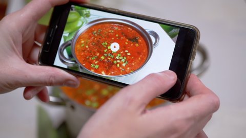 A Woman Looks at a Photo of Traditional Ukrainian Red Borscht on a Smartphone. Photo of food through the camera of a mobile phone in a mobile application. Home kitchen. Lunchtime. Fingerprints.