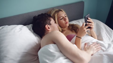 Nice girl staring mobile phone in bed. Sad woman and man, husband in bed, lack of sexual activity in family. Young unhappy couple problem in bedroom, quarrel between lovers. Divorce, marriage
