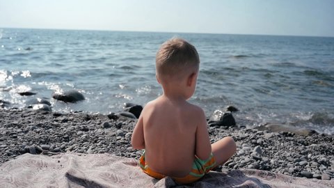 A boy-child sits with his back to the camera and throws a small pebble into the sea. Vacation on the seashore with the whole family.