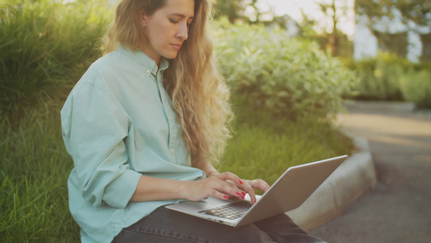 Portrait of young beautiful female freelancer sitting on border stone on street, typing on laptop and then smiling at camera