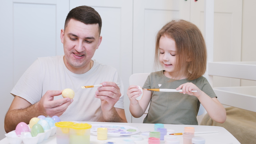 Happy easter and happy family. Father and daughter sitting at white table at home and painting eggs with gouache while preparing for Easter holiday.