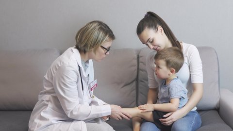 pediatrician doctor visits baby boy child at home. caucasian kid sitting mother's knees grey sofa living-room. female woman doctor in white uniform, examines toddler's leg and knee, asking symptoms 