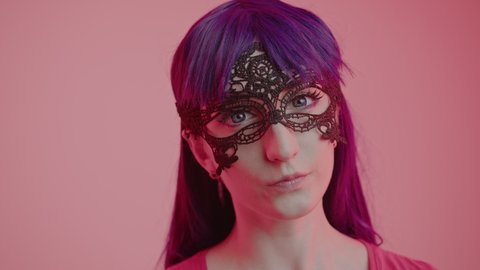 Mysterious Caucasian Woman in a lace mask looking intensively into the camera close-up shot pink background. High quality 4k footage