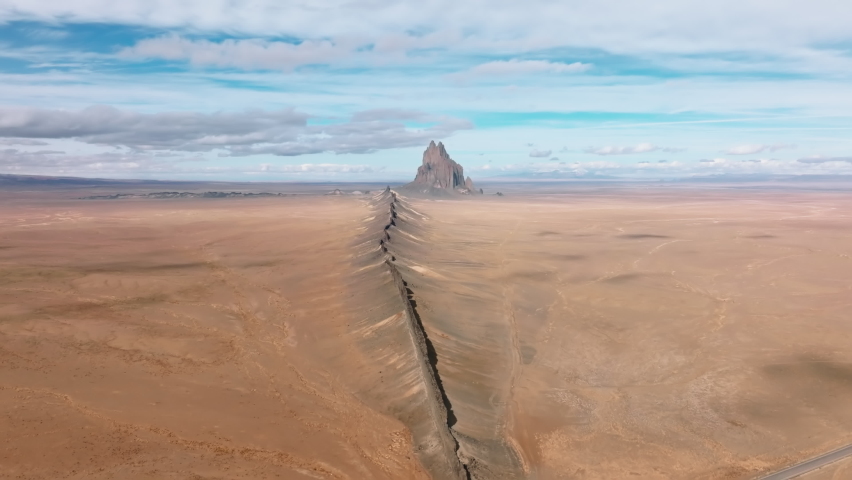 Unique terrain without any greenery below cloudy blue skyline. Drone footage of an enormous crack towards Shiprock formation in the middle of the desert. High quality 4k footage Royalty-Free Stock Footage #1087792947