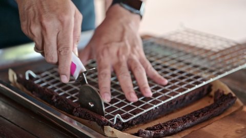Baker hand using wheel cutter cutting on baked dark chocolate chip brownies in tray on wooden table in bakery shop