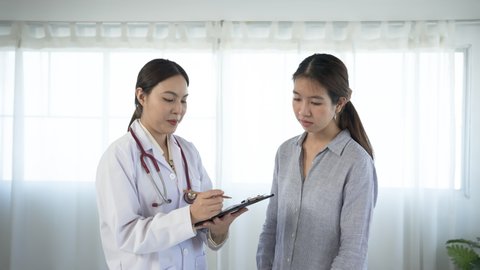 4K 50fps, white Asian female doctor standing and taking notes on a long-haired Asian patient wearing a long-sleeved shirt in a hospital examination room..	
