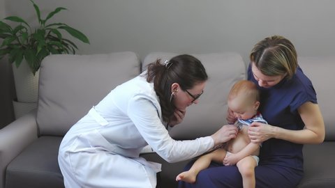 home appointment woman pediatrician child baby and mother, doctor in uniform examines with stethoscope kid's lungs, listens heartbeat, mother helping doing checkup. living room, grey sofa, boy son 