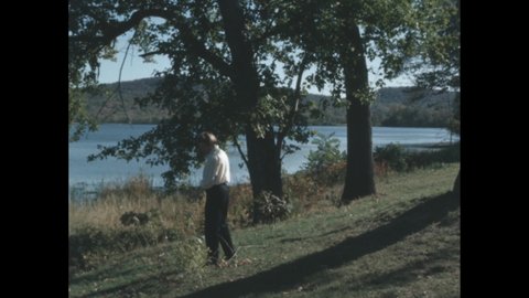 1960s: Man standing lakeside. Hand grabbing flowers. Person grabbing plant in tall grass.