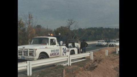1960s: Person operating tow truck on roadside. Person lifting car onto tow truck. Police cars blocking highway. Trees, plants lining lake edge. Lake shoreline.
