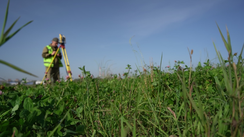 The surveyor in the background is at work measuring distances. In the foreground the green grass is in focus. Royalty-Free Stock Footage #1087812963