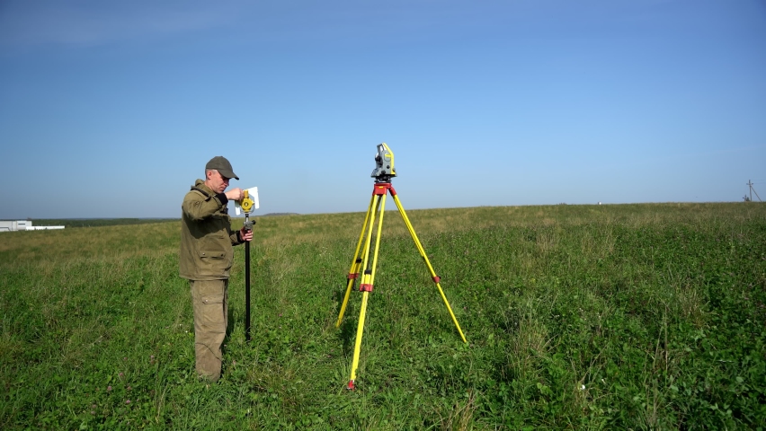 
The surveyor with the reflector in his hand prepares to take measurements near the surveying equipment. 
Surveyors ensure precise measurements before undertaking large construction projects. Royalty-Free Stock Footage #1087812965