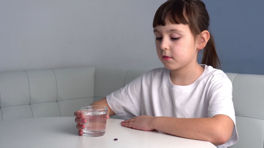 Cute child in T-shirt takes medecine and drinking glass of water. Little girl takes pill or vitamin