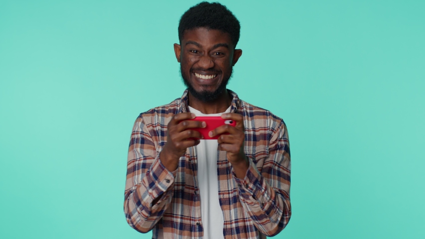 Worried funny addicted african american man enthusiastically playing racing video games on mobile phone. Young guy using smartphone gadget app with drive simulator isolayed on blue studio background