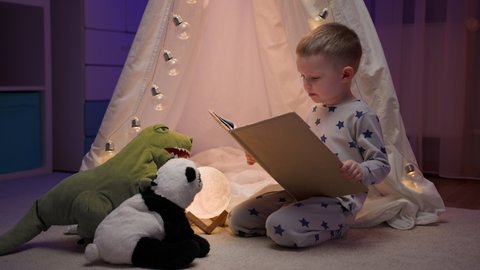 Blond boy in pajamas reading fairy tale to his stuffed panda toy and green dinosaur sitting comfortably next to tent in dark night bedroom. Love concept, childhood, friendship, imagination, creativity
