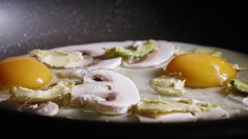 Salt, Black Pepper, And Cayenne Pepper Sprinkling On Eggs With Mushroom And Avocado Cooking In A Frying Pan. close up