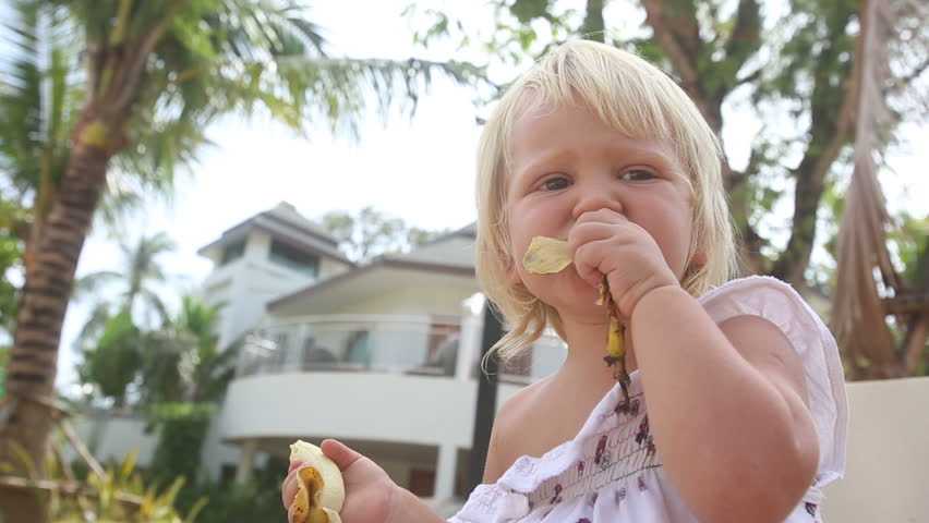 blonde little girl gives back banana skin to mother near swimming pool against hotel buildings