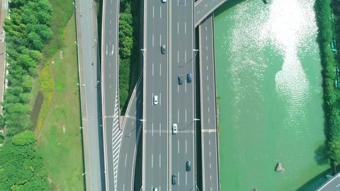 Aerial top down view of a highway overpass multilevel junction with fast moving cars surrounded by green trees and with a river on a side on a sunny day