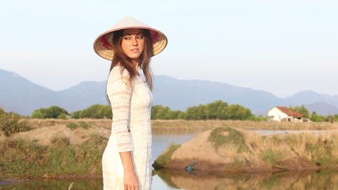 european blonde girl in vietnamese national costume and hat poses near pond for paddy planting against mountains