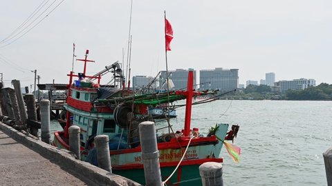 Pattaya , Chonburi , Thailand - 12 03 2021: A fishing boat docked with a red flag while a motorcycle passes by on the left and the city of Pattaya at the background, Pattaya Fishing Dock, Chonburi, Th