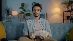 Portrait of pensive Middle Eastern man watching TV eating popcorn and thinking in dark apartment in the evening. Mass media and emotion concept.