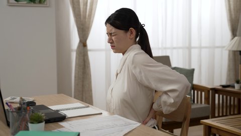 side view of a stressed asian female entrepreneur having painful backache is pounding massaging her lower back while working from home at desk