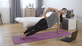 tanned sportive woman is watching a fitness video on the internet with her notebook while practicing side plank core exercise in the morning at home.