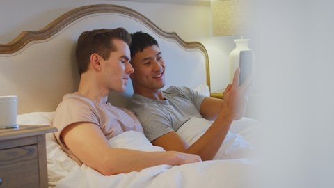 Loving same sex male couple lying in bed at home making video call on mobile phone - shot in slow motion