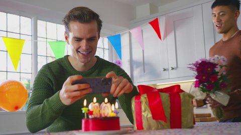 Same sex male couple celebrating 30th birthday at home with man taking photo of cake on phone as partner gives him flowers - shot in slow motion