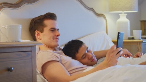Loving same sex male couple lying in bed at home with man taking selfie on mobile phone whilst partner sleeps - shot in slow motion