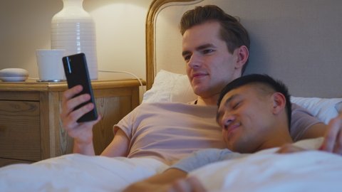 Loving same sex male couple lying in bed at home with man looking at social media on mobile phone whilst partner sleeps - shot in slow motion